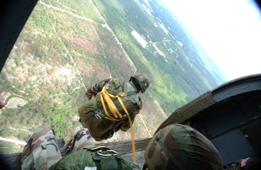 View from inside a military aircraft, paratrooper preparing to exit the door over forested terrain.